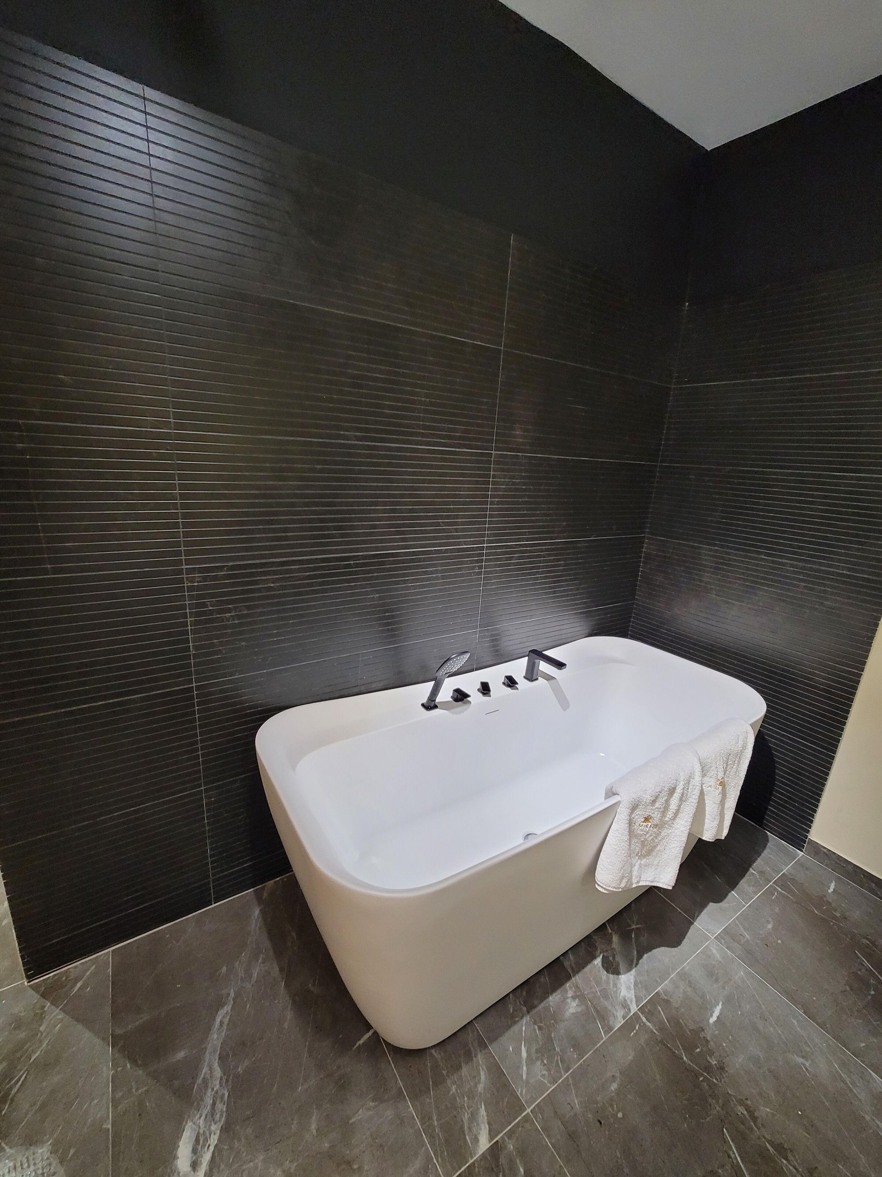 A modern bathroom with a freestanding bathtub, dark tiled walls, and a marble floor.