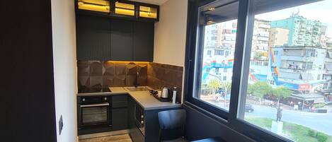 A modern kitchen with dark cabinetry, a marble countertop, and a dining area with a table and chairs. A window provides a view of the cityscape.