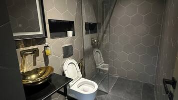 Un baño moderno con ducha envuelta en vidrio, un lavamanos dorado y un inodoro con tazón blanco.