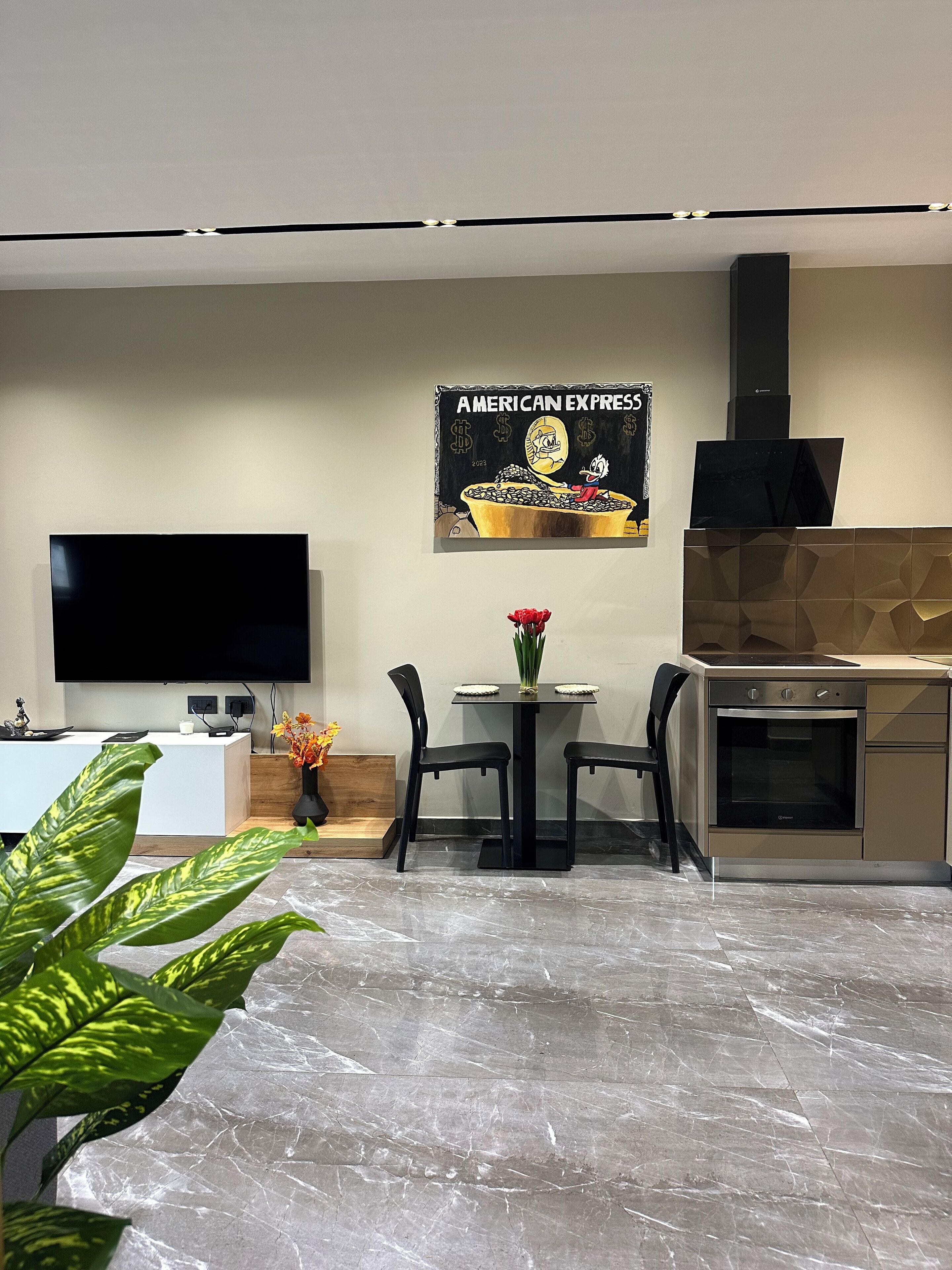 A modern kitchen with a built-in oven, a dining table with chairs, a flat-screen TV, and a wall-mounted sign.