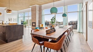 Un vaste espace dédié à la salle à manger, avec une longue table en bois et des chaises en cuir, une cheminée en briques et de grandes fenêtres offrant une vue sur l’extérieur.
