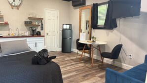 A compact room with a bed, a small dining table with chairs, a refrigerator, a clock, and a window with curtains.