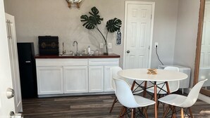 A dining area with a round table and white chairs, a kitchen with white cabinets, a clock on the wall, and a ceiling fan.