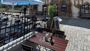 An outdoor seating area with tables and chairs, a large ornate building in the background, and a clear blue sky with clouds. - New! Modern Bavarian flair with Mountain View in front of famous Pilatushaus! (Oberammergau)