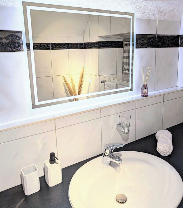 A bathroom with a large mirror, a sink, a glass, a bottle of soap, and a towel.
