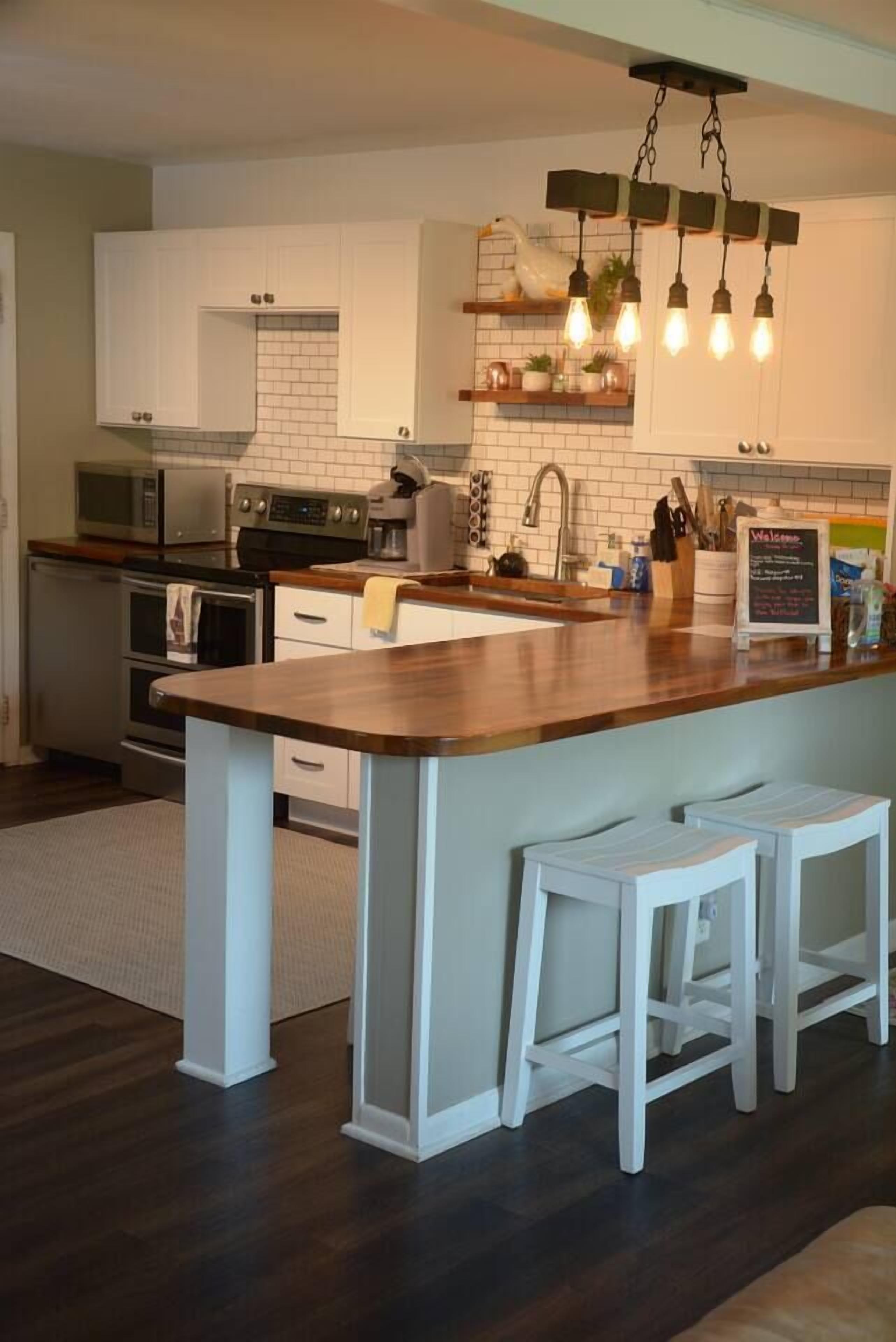 Una cocina con armarios blancos, una isla de madera con taburetes, una lámpara colgante y un letrero en una pizarra.