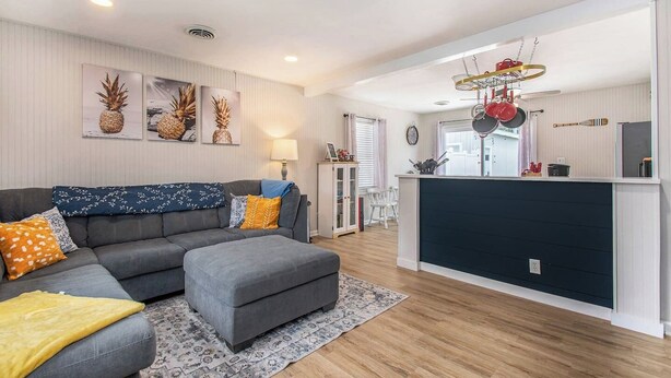 A modern living room with a grey sofa, a small ottoman, and a patterned rug. There is a kitchen area with a dark countertop and hanging pots.
