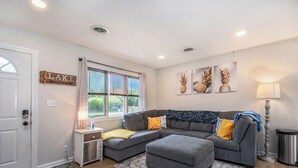 A modern living room with a grey sectional sofa, a small side table, a lamp, and a 'LAKE' sign on the wall.