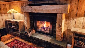 Ein gemütlicher Kamin mit brennenden Holzscheiten, Holzregale und eine Lampe.