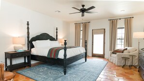 A bedroom with a four-poster bed, a ceiling fan, a nightstand with a lamp, a chair, a dresser, and a window with curtains.