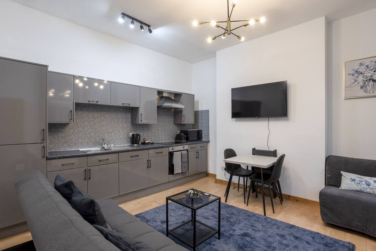 A modern kitchen with grey cabinets, a dining area with a table and chairs, a grey sofa, and a flat-screen TV mounted on the wall.