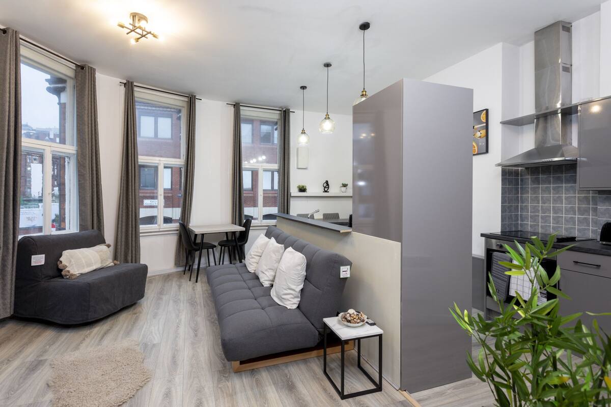 A modern living room with a grey sofa, a small table with a bowl, and a kitchen area with grey cabinets and a stove.