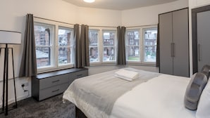 Une chambre avec un grand lit, un bureau et une armoire. De nombreuses fenêtres offrent une vue sur les bâtiments environnants.