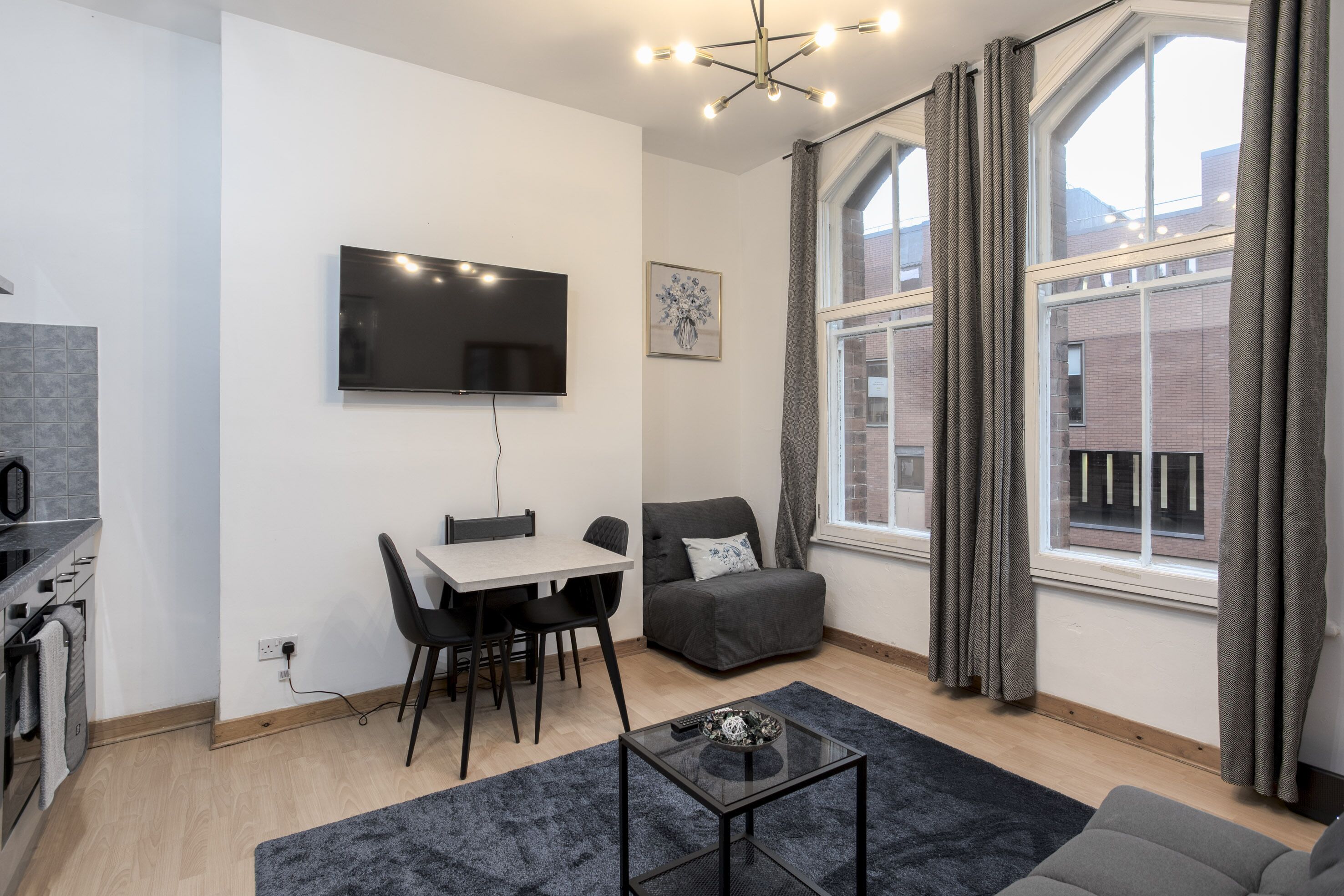A modern living room with a flat-screen TV, a dining table with chairs, a gray sofa, and a large window with curtains.