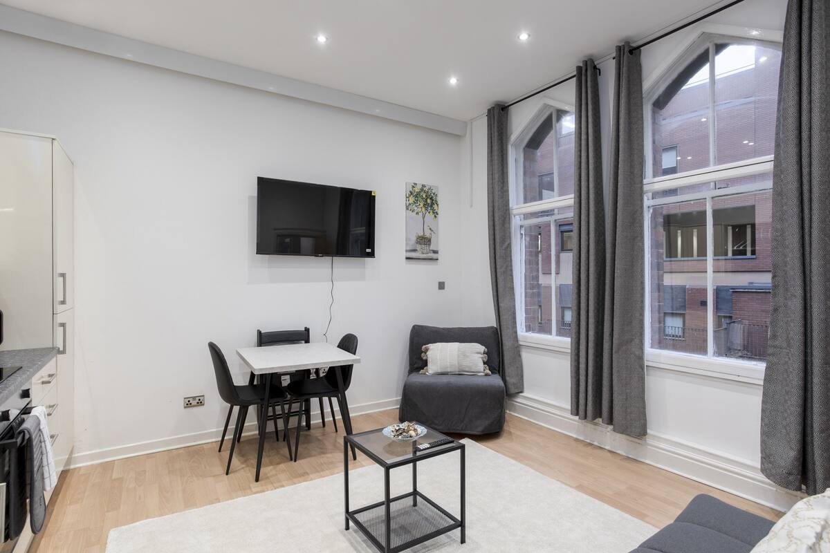 A modern living room with a dining area, a flat-screen TV mounted on the wall, a grey armchair, and a large window with curtains.