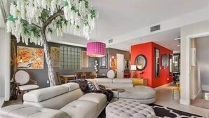 A modern living room with a large sofa, a round ottoman, and a tree with white flowers. There is a red accent wall and a black and white rug. - Little Harbor Resort at Bahia Beach (Ruskin)