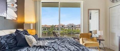 A bedroom with a large bed, a chair, a lamp, a clock, and a view of a balcony with houses.