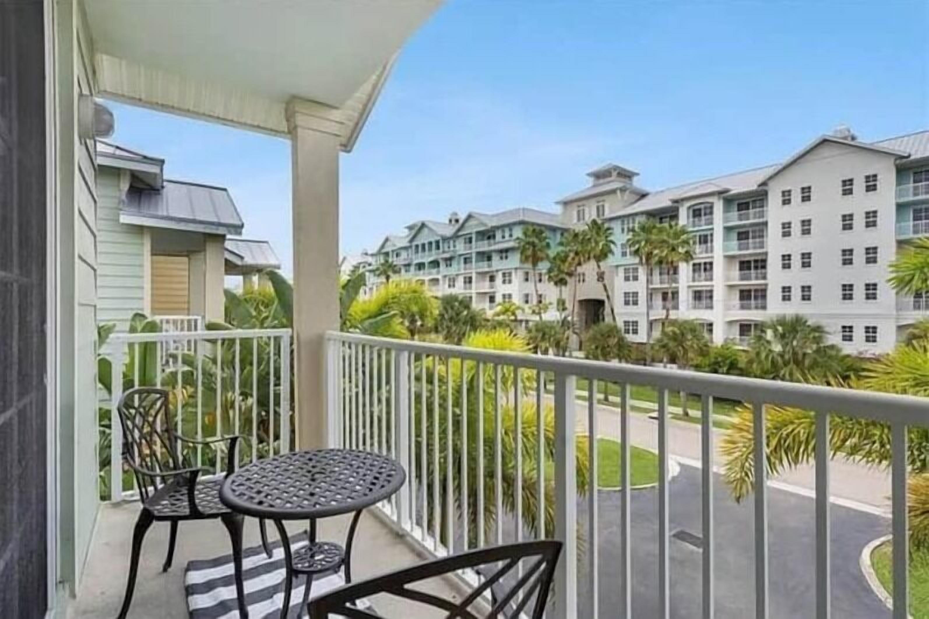 A balcony with a table and chairs, overlooking a parking area and a building with multiple floors and balconies.