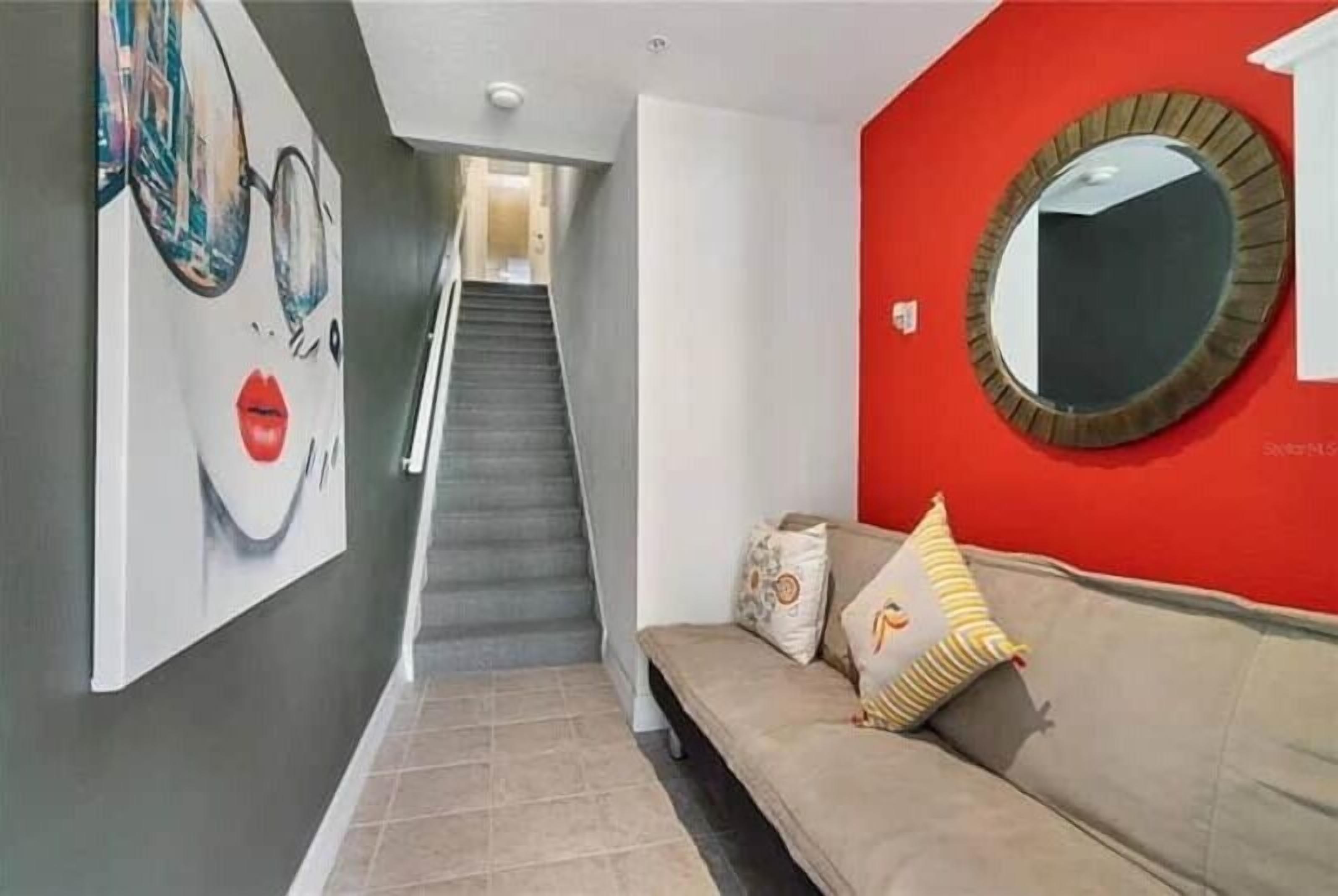 A hallway with a staircase, a red accent wall, a round mirror, and a sofa with cushions.