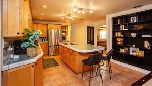 A kitchen with wooden cabinets, a stainless steel refrigerator, a large island with bar stools, and a bookshelf with various items. - New! Saltwater pool home with hot tub, game room, and outdoor games! (Gainesville)
