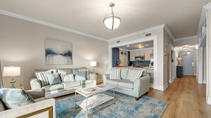 Un salon moderne avec deux canapés, une table basse et un tapis. On aperçoit une cuisine à l’arrière-plan, avec un coin repas.