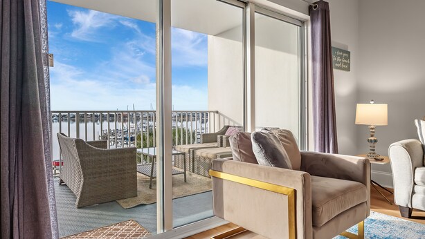 Un salon moderne doté d’une grande porte-fenêtre coulissante donnant sur un balcon avec vue sur un port de plaisance. On y trouve deux canapés et une petite table avec une lampe.