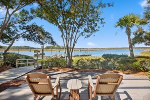 A patio with two wooden chairs and a table, overlooking a lake with a dock and a signpost. - Modern Riverfront Inlet Cove 3Bd- Open Porches, Dog Friendly, Pool Access! (Kiawah Island)