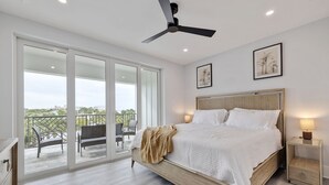 Une chambre avec un grand lit, un ventilateur de plafond, une table de chevet avec une lampe et un balcon avec des chaises.