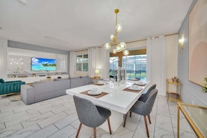 Una sala moderna con un televisor de pantalla plana grande, una zona de comedor con una mesa blanca y sillas grises, y un sofá gris.