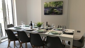 Een eethoek met een witte tafel gedekt voor zes personen, zwarte stoelen, een potplant en een schilderij van een boslandschap.