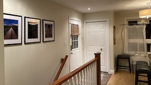 Un couloir avec des tableaux encadrés accrochés au mur, une porte blanche, une fenêtre avec un rideau et un escalier avec une rampe en bois.