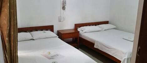 A room with two single beds, each with a wooden headboard and white bedding. There is a wooden nightstand between the beds, and a wall-mounted electrical outlet above it.