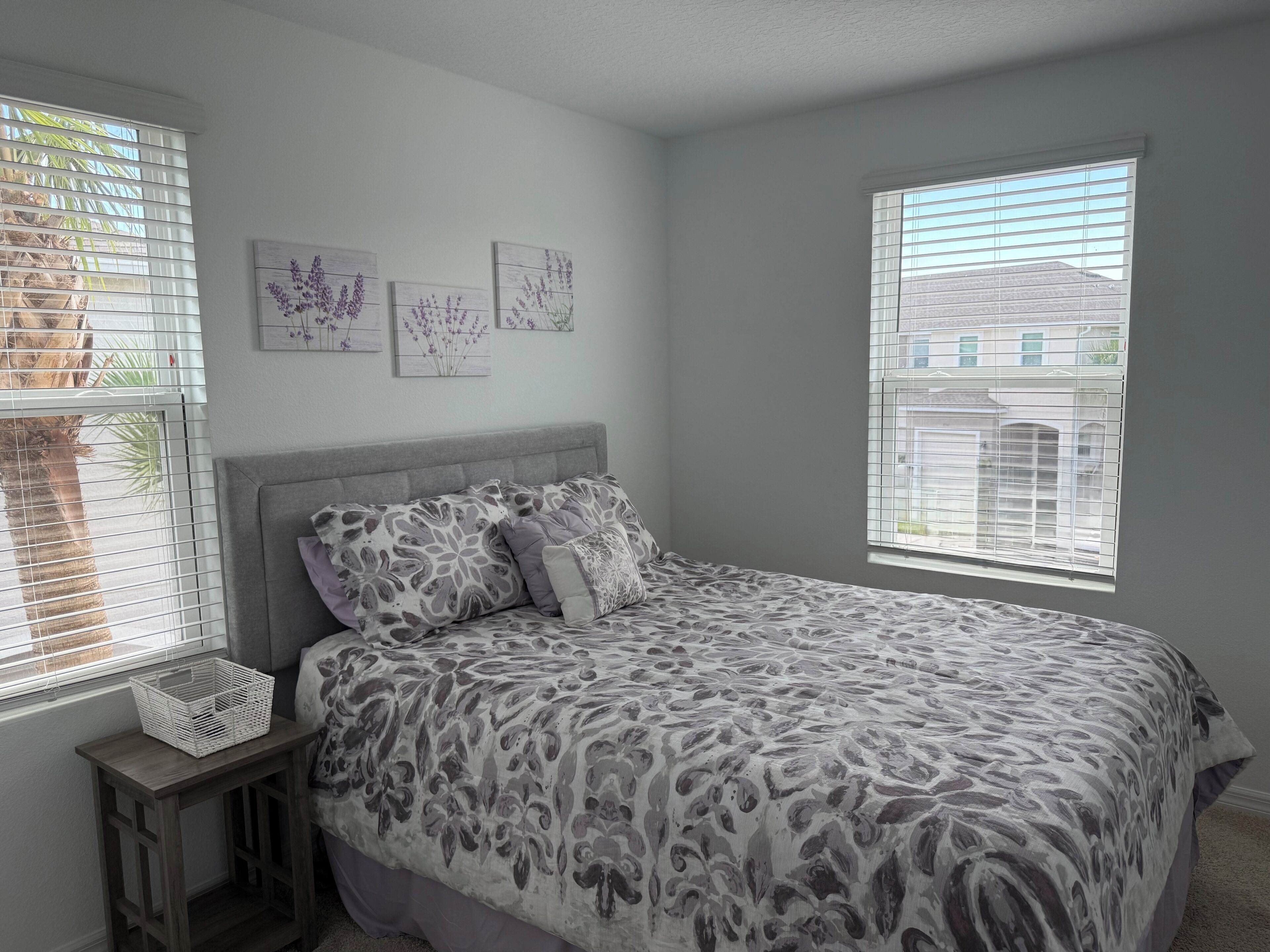A bedroom with a bed, two windows, a small table, and wall art.