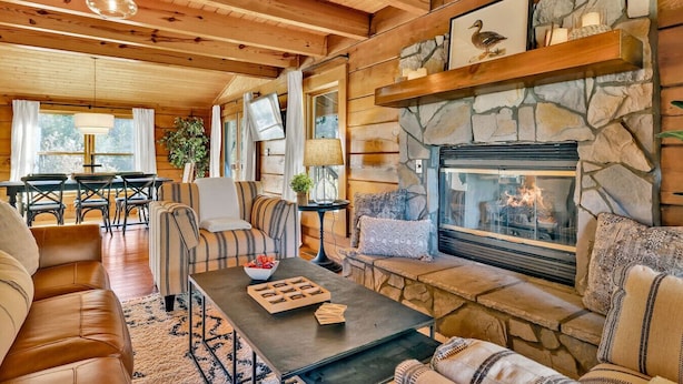 A cozy log cabin interior with a stone fireplace, wooden furniture, and a dining area.