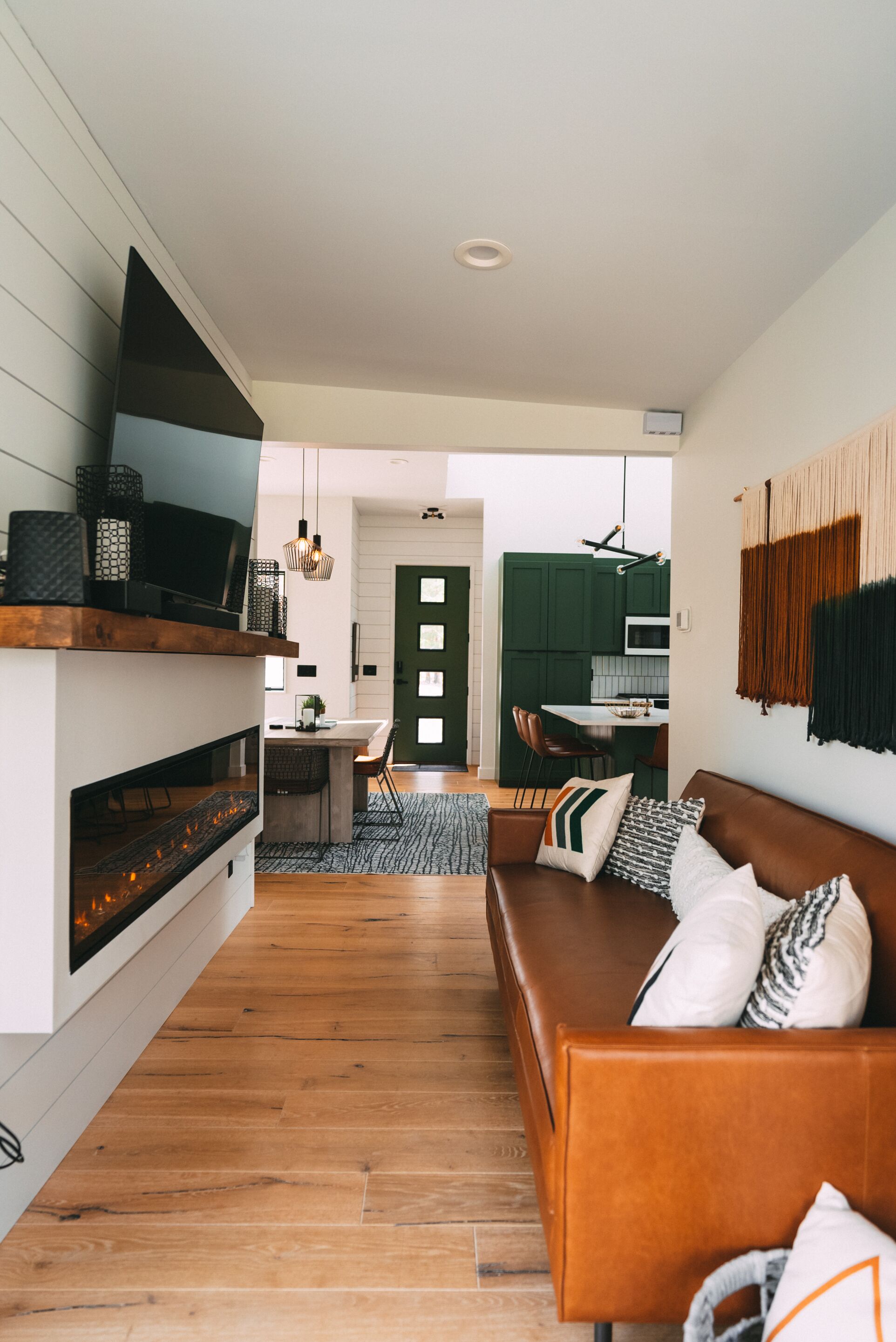 A modern living room with a wooden floor, a brown leather sofa, and a flat-screen TV mounted on a white wall.