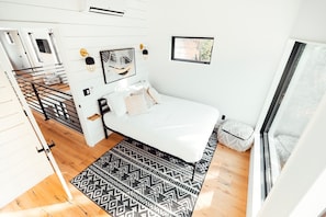 A modern bedroom with a white bed, patterned rug, and a view of the outdoors through a window.