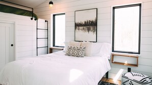 Un dormitorio con cama, ventilador de techo, dos lámparas colgantes, una ventana y un cuadro en la pared.