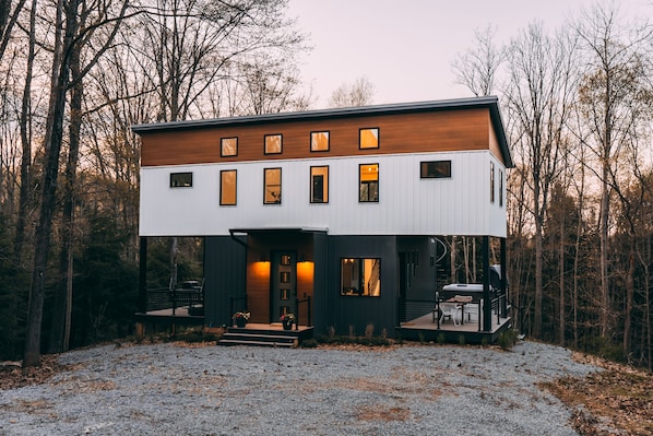 A modern two-story cabin with a wooden upper floor and a white lower floor, surrounded by trees.