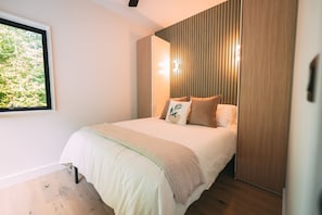 A modern bedroom with a large bed, a window with a view of greenery, and a wooden panel wall.