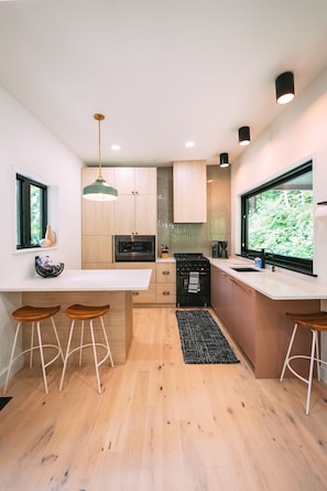 A modern kitchen with wooden cabinets, a central island with stools, a built-in oven, and a large window offering a view of greenery.