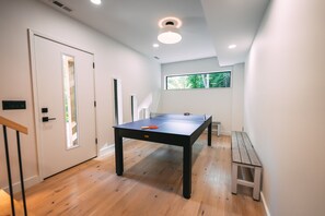 A room with a ping pong table, a bench, a door, and a window with a view of greenery.