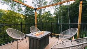 Een terras buiten met een rechthoekige vuurplaats, twee rieten stoelen en een houten balustrade.