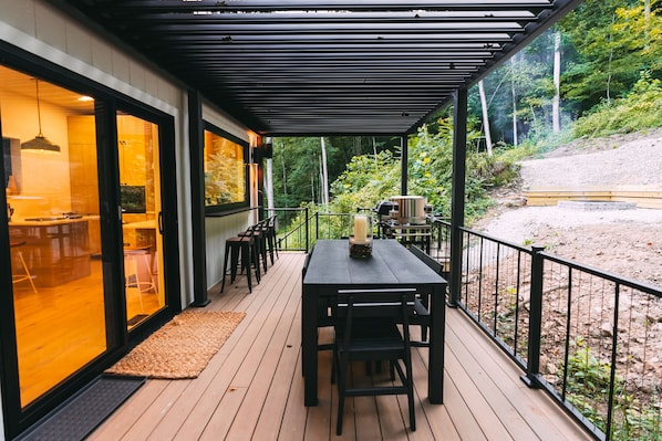 A wooden deck with a table and chairs, a pergola, and a view of a forested area.