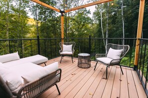 A wooden deck with two lounge chairs and a small table, surrounded by a black railing and overlooking a forested area.