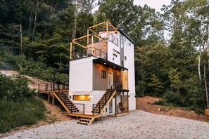 A modern multi-level cabin with a staircase leading to a balcony, surrounded by a forested area.