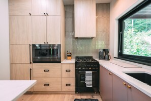 A modern kitchen with wooden cabinets, a gas stove, an oven, and a window with a view of greenery.