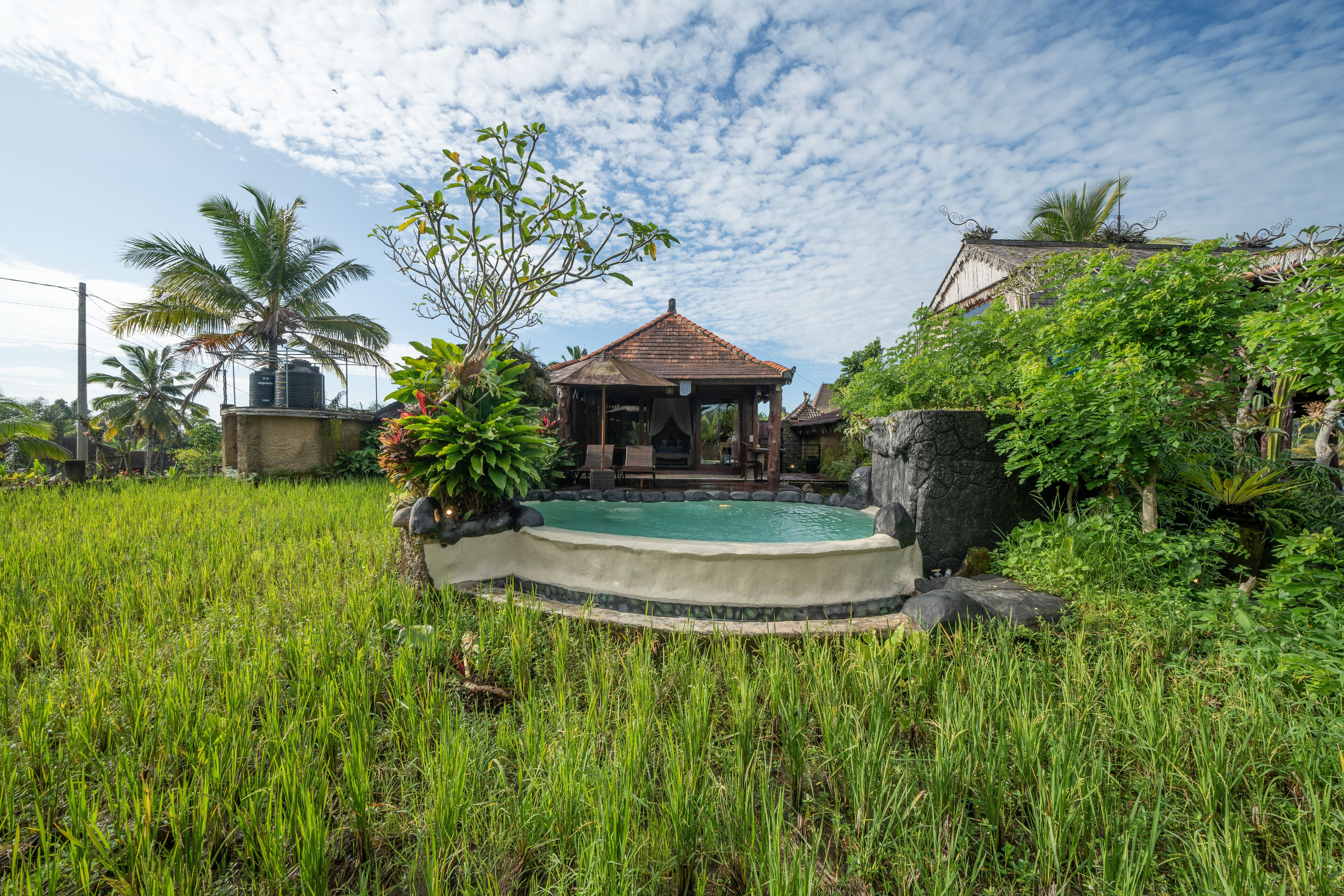 A traditional wooden gazebo with a tiled roof, surrounded by lush greenery and a small pool.