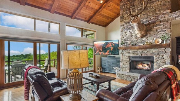 A spacious living room with leather sofas, a stone fireplace, a mounted deer head, and a large TV displaying a mountain scene. - Spacious Home on Beech Mountain w/ Views (Beech Mountain)