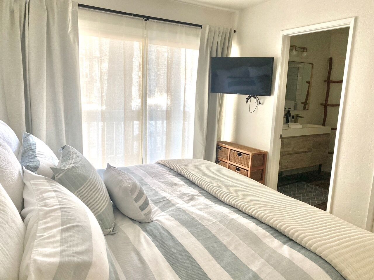 A bedroom with a bed, curtains, a television, a wooden dresser, and a bathroom visible through an open door.