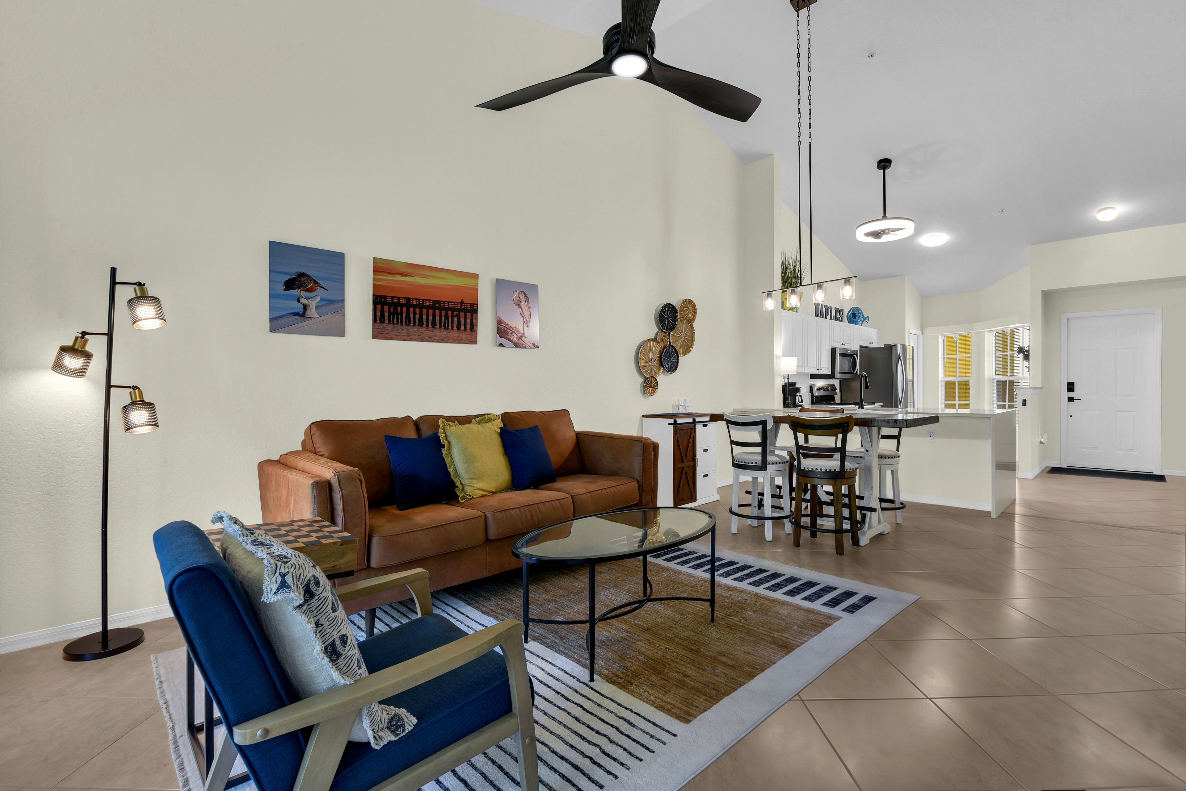 A spacious living area with a brown leather sofa, a glass coffee table, and a blue armchair. There is a kitchen in the background with white cabinets and a dining area.